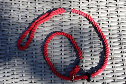 A cotton raspberry red slip lead with a rubber stopper and bindings on a grey rattan chair. Loosely spiralled.