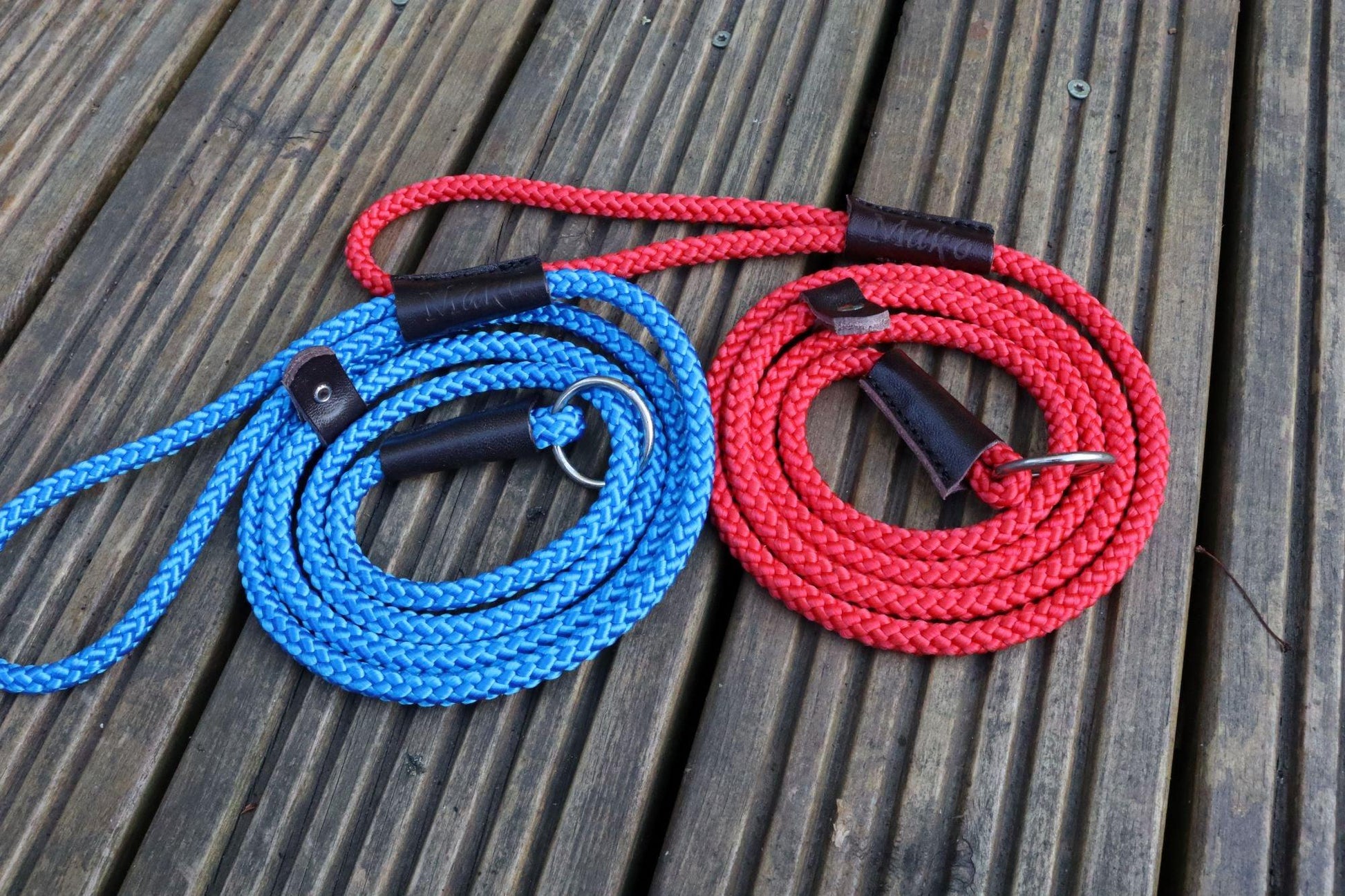 One red and one blue slip leads on wooden decking. Both leads are tightly spiralled with the handle pointing left and demonstrating the embossed Mako stamp in the leather binding.